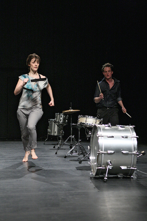 A woman runs alongside a drum kit that's arranged in a straight line while its drummer stands on the other side.